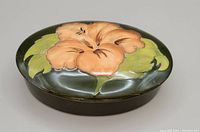 Oval Moorcroft pottery covered dish, dark green background with coral hibiscus flower on lid, top view.