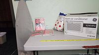 GE 5150 BTU room air conditioner in box next to ironing board cover and pad on table with measuring tape.