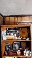 Overall view of wooden shelving unit displaying art supplies including paint sets, brushes, and paper.