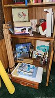 Photo showing LaminatorIt laminator box, white paper cutter, wooden easel, paint brushes, watercolor paint sets, art books, and stacked paper on wooden shelf.