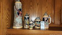 Photo of five beer steins of varying sizes lined up on a wood shelf, showing full set and lid styles.