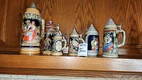 Photo showing all six beer steins lined up on a wooden shelf, varying in size and detailed relief decoration with metal lids