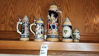 Photo showing five beer steins in a row on a wooden shelf, varying in height and design, mostly ceramic with traditional German motifs and pewter lids.