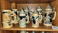 Front view of six German-style ceramic beer steins with metal lids on wooden shelf, showing detailed colorful designs and motifs.
