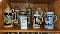 Front and side views of six German beer steins placed on wooden shelves showing variety in designs and colors.