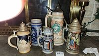 Wide view showing all five German-style ceramic beer steins with pewter lids, varying sizes around fireplace area, some colorful relief decoration visible.