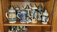 Front view of 7 ceramic beer steins with pewter lids on wood shelf, showing different painted and relief scenes