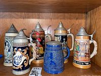 All 7 beer steins shown on wooden shelf, front view showing designs, lids, and tags