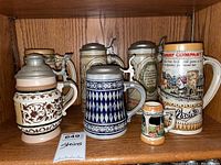 Photo showing six beer steins arranged on wooden shelf, details of different sizes and designs visible.