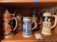 Photo of 5 decorative German beer steins with metal hinged lids placed on a wooden shelf, showing various traditional motifs and relief work.