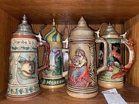 Four detailed ceramic beer steins with ornate metal lids and colorful relief artwork representing different themes and locations.