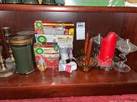 Photo showing various gold taper candle holders, a green scented candle with wooden lid, red pillar candle, brown taper candles, and Air Wick Essential Oils air fresheners and refills in original packaging.