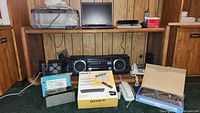 Photo showing a Fisher boombox with dual speakers on either side, Sony WebTV box, cordless phones, remotes, and an electronic component under a wooden desk.
