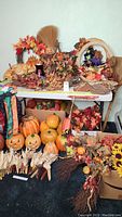 Wide view of assorted fall and Halloween decor displayed on table and floor: artificial pumpkins, foliage wreaths, corn stalks, baskets, and small pumpkins in boxes.
