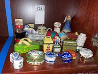 Shelved trinket boxes various shapes and colors including handbags, hexagonal, round, figurines, and buildings in porcelain and ceramic.