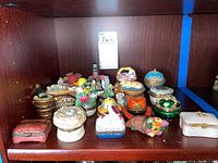 Photo showing a shelf with multiple small hinged trinket boxes of various shapes, sizes, and colors, including animal, fruit and basket shaped ones