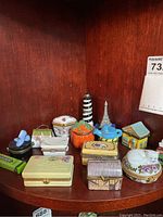 Photo of multiple hinged trinket boxes in various shapes, sizes, and styles displayed on wooden shelf with a mix of decorative motifs including animals, buildings, and novelty shapes.