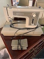 Front view of beige Kenmore portable sewing machine sitting on matching beige hard detachable cover, with power cord and pedals placed in front.