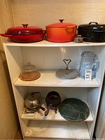Red, orange, and black Dutch oven style pots on top shelf, glass pitcher, small metal bowl with lid, metal kettle, small metal pot with handle, and metal lid on lower shelves.