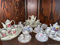 Overview of four ornate miniature tea sets with porcelain teapots, cups, saucers, creamers, and sugar bowls in floral and leafy designs with gold trim, arranged on shelves.