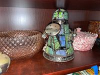 Shelf display showing stained glass and metal watering can, pink diamond-patterned glass bowl, and a pink lidded ceramic dish