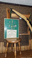 Wooden wall hanging sportsman shelf mounted on the wall with a framed certificate and metallic pins placed on the shelf, wooden chair in front, leather carrying case leaning against the chair.