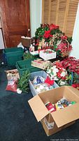 Wide angle photo showing multiple green bins filled with Christmas decorations, several wreaths with red bows, poinsettias, nativity scene figures, and assorted holiday decor arranged on dark carpet.