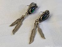 Pair of sterling silver earrings with green oval stones and feather-shaped dangles shown on neutral fabric background