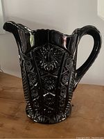 Side view of vintage black Tiara glass pitcher showing monarch pattern, scalloped top, curved handle, and spout.
