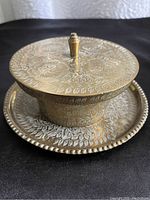 Front angle view of the smaller brass lidded trumpet bowl showing floral etching on lid and base plate with raised beaded edge.