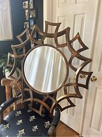 Mirror shown leaning against a door with a chair nearby, highlighting the frame's rustic bronze geometric sunburst design and reflective round center.