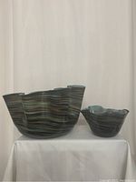 Front view of two ruffle edged hand blown glass bowls with swirling blue and green hues on white cloth and curtain background.