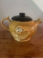 Front view showing the yellow ceramic teapot with the whimsical face detail and black lid.
