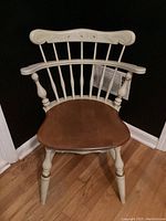 Front view of the vintage Ethan Allen chair showing white and gold painted comb back with a wooden seat on a wood floor against black walls.
