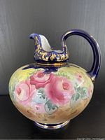 Front side of the vintage hand painted Japanese pitcher showing pink roses and gold detail on cobalt blue handle and spout.
