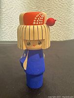Front view of a wooden geisha girl figure painted with blue kimono and red headpiece detail.