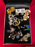 Overview of assorted jewelry pieces including earrings, pendants, rings, brooches on black display inside red box