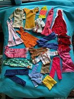 Full spread of assorted vintage Barbie doll clothes showing various styles and colors arranged on teal background.