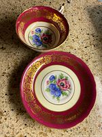 Overhead view of cup and saucer showing hand painted blue and pink flowers in center, gold ornate pattern on pink background.