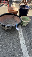 Photo showing four garden pots of various sizes and finishes, placed on asphalt. Includes a large black glazed pot with wear, a tall dark blue glazed pot, a green pot with a chip, and a terra-cotta pot.