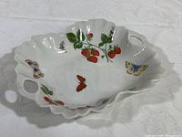 Top front view of the scalloped white Limoges candy dish adorned with painted strawberries and colorful butterflies. Dish has two handles on opposite sides.