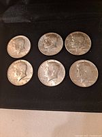 Obverse side of six Kennedy Half Dollar coins arranged on a black surface, showing dates 1965, 1966, and 1967 with wear and tarnish.