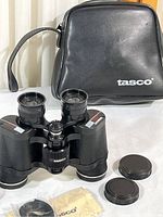 Tasco binoculars and accessories including vinyl pouch, lens caps, strap, and cleaning cloth arranged on a white cloth surface.