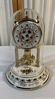 Front view of vintage anniversary clock displaying white porcelain dial with Roman numerals, gold hands, floral decorations, gold-tone rotating pendulum balls, and clear glass dome.