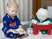Side angle showing both Japanese bisque student figurines: girl reading a book and boy writing at desk, with box labeled 'Japanese Gump’s Study Hard Boy & Girl' in background.