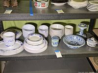 Photo showing a mix of blue and white dinnerware including plastic plates and bowls, ceramic small jars, a covered dish, and a candlestick holder.