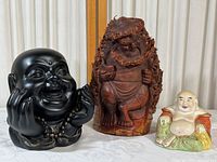 Three Buddha figures displayed together on a white cloth background showing all three statues clearly: black composite bust, carved wood figure, and painted ceramic seated statue.