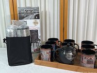 Photo showing travel mug set with two stainless steel mugs in black carrying bag next to box, and bamboo tray holding seven Egyptian motif coffee mugs and small glass tea carafe with black handle and lid.