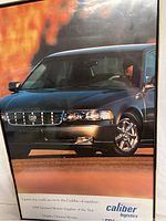 Close-up of the front side of the dark-colored Cadillac car with driver visible through the window, set against orange background on the poster.