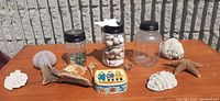 Three vintage glass jars with black lids, two filled with sea shells, one empty, alongside assorted sea-themed decorative items including coral pieces, starfish, sea urchin shell, and a decorative metal tin. All items arranged on a wooden table outside against a gray brick wall.